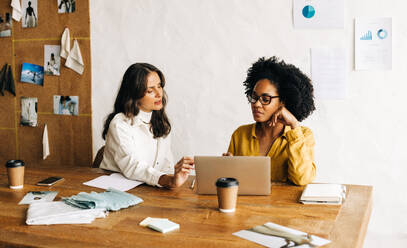 Geschäftsfrauen, eine Modedesignerin und ihre Kollegin, nutzen einen Laptop, um in ihrem Büro kreativ über Stoffdesigns zu diskutieren. Unternehmerinnen beim Brainstorming über Lösungen für ihr Bekleidungs-Startup. - JLPSF30149