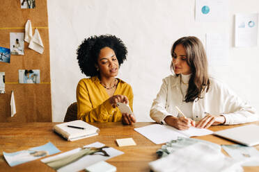 Kreative Geschäftsfrauen, beide Modedesignerinnen, sitzen zusammen in einem Büro und diskutieren kreative Ideen mit Hilfe eines Smartphones. Zwei Bekleidungsdesignerinnen nutzen ihre unternehmerischen Fähigkeiten, um ihr Start-up voranzubringen. - JLPSF30150