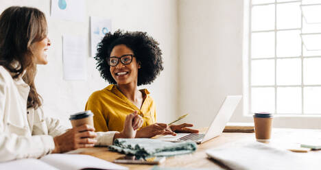 Professional business women sit together in an office, brainstorming creative ideas for their fashion design business. Young clothing designers work towards achieving their entrepreneurial goals. - JLPSF30153