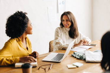 Female team of business professionals, including a fashion designer and entrepreneur, are happily working together in an office to plan for the success of their small business. - JLPSF30155