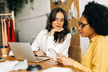 Ein Team von Geschäftsfrauen arbeitet mit Hilfe eines Smartphones und eines Laptops im Büro an einem Modedesignprojekt. Bekleidungsdesignerinnen arbeiten zusammen und nutzen ihre Kreativität für ein erfolgreiches Unternehmertum. - JLPSF30159