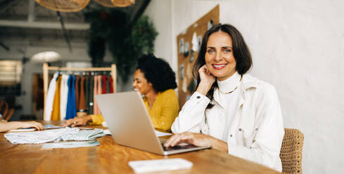 Eine Bekleidungsdesignerin lächelt in die Kamera, während sie in einem Modegeschäft an einem Laptop arbeitet. Eine Geschäftsfrau arbeitet in einer Boutique mit ihren Kolleginnen. - JLPSF30164