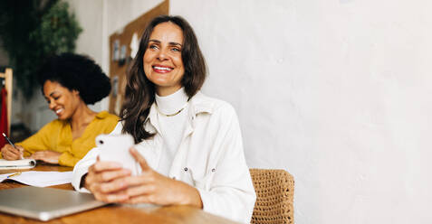 Female entrepreneur smiling and looking away thoughtfully in a clothing design office. Business woman using a smartphone while sitting at her desk. - JLPSF30167