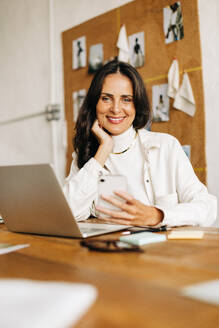 Eine kreative Modedesignerin und Geschäftsfrau sitzt in ihrem Büro, lächelt und schaut selbstbewusst in die Kamera. Eine glückliche Geschäftsfrau, die ein Smartphone und einen Laptop benutzt, um ein kleines Design-Startup zu leiten. - JLPSF30170