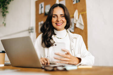 Small business owner sits in her office, using a laptop and mobile phone while smiling happily. She is an entrepreneur and fabric designer who loves being creative with fashion design. - JLPSF30171