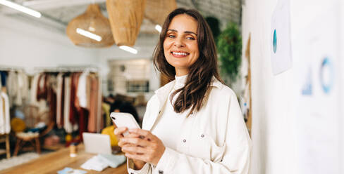 Portrait of a female fashion designer using a smartphone in a clothing boutique. Woman using mobile technology for text messaging making schedules and contacting clients in a small business. - JLPSF30189