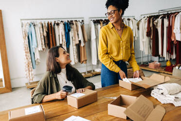 Erfolgreiche Geschäftsfrauen lächeln sich an, während sie Bestellungen in ihrem Online-Kleiderladen erfüllen. Glückliche Unternehmerinnen, die als Team ein Dropshipping-Geschäft betreiben. - JLPSF30197