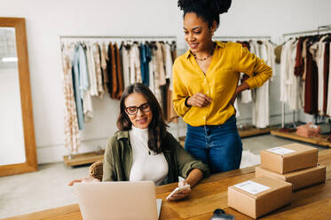 Frauen, die an einem Laptop die Zahlen und Fortschritte ihrer E-Commerce-Website besprechen. Zwei Unternehmerinnen, die ein erfolgreiches Bekleidungsgeschäft betreiben, das Bestellungen per Dropshipping abwickelt - JLPSF30199