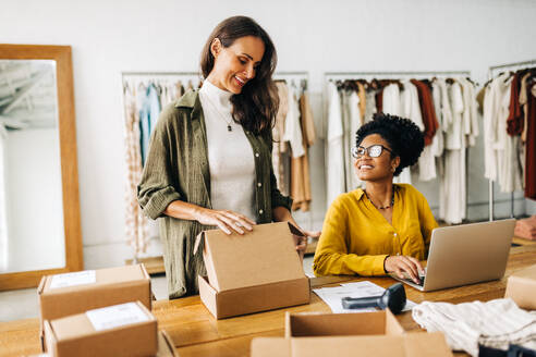 Two happy business women working as a team in fulfilling orders for their online clothing store. Female entrepreneurs venturing into dropshipping as they expand their boutique with an ecommerce site. - JLPSF30207