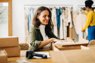 Eine Geschäftsfrau arbeitet glücklich in ihrem Boutique-Bekleidungsgeschäft und bereitet ein Paket für den Versand vor, um ihr erfolgreiches Online-Geschäft zu starten. - JLPSF30216