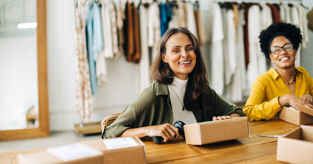 Focused on retail success and growth, two business women expand their small business with an online store and dropshipping services. Women smiling at the camera as they fulfil clothing orders. - JLPSF30222