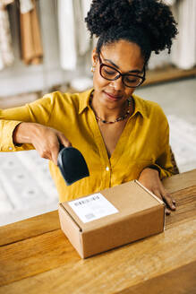 Black female entrepreneur using a barcode scanner to scan a QR code as she processes a package for dropshipping. Young business woman using her ecommerce skills to run a reliable online store. - JLPSF30224