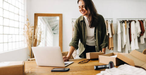 Successful female entrepreneur using a laptop to manage her online store and process shipping orders on an ecommerce platform. Happy business woman fulfilling clothing orders in her boutique. - JLPSF30232