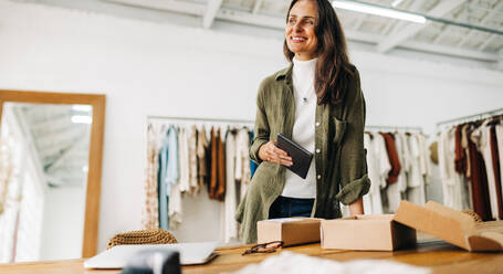 Die Inhaberin eines Kleinunternehmens steht in ihrem Laden und lächelt, als sie über das Wachstum und den Erfolg ihres Online-Shops nachdenkt. Eine Frau, die eine E-Commerce-Boutique betreibt, verwendet ein Tablet, um die Bestellungen abzuwickeln. - JLPSF30233