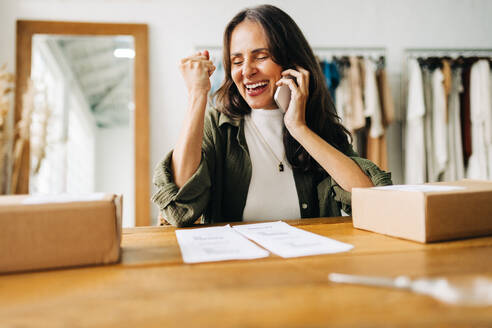 Business woman achieves success as an ecommerce entrepreneur, she celebrates as she receives a phone call from her shipping team. Female clothing store owner running an online retail business. - JLPSF30236