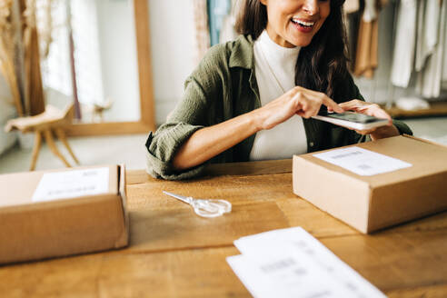 Happy business woman processing clothing orders for her ecommerce store, scanning a QR code with her mobile phone in order to track the package during the dropshipping process. - JLPSF30237