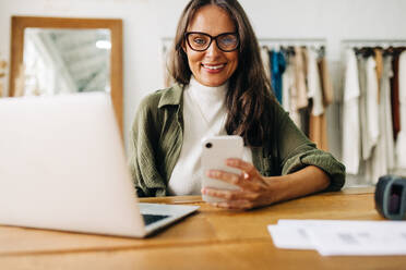 Eine Frau lächelt in die Kamera, während sie ihr Mobiltelefon benutzt, um ihr kleines Unternehmen effizient zu koordinieren. Eine glückliche Unternehmerin, die eine E-Commerce-Website für ihre Bekleidungsboutique einrichtet. - JLPSF30244