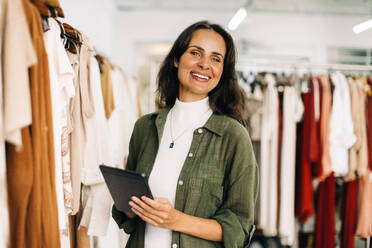 Successful boutique owner is standing in her store, her tablet in hand, as she manages her business operations with ease. Happy business woman taking inventory for her small business. - JLPSF30246