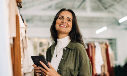 Kleinunternehmerin, die ihre Bekleidungsboutique leitet und ein Tablet in der Hand hält, während sie ihre täglichen Aufgaben erledigt. Zufriedene Unternehmerin, die durch den Bestand und die Bestellungen ihres Geschäfts navigiert. - JLPSF30247