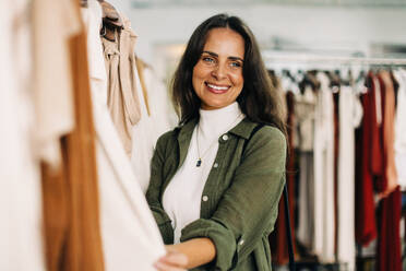 Eine Kundin stöbert in einem Modegeschäft durch die Kleiderständer und wählt sorgfältig aus, welche Artikel sie kaufen möchte. Eine Frau lächelt, während sie nach hochwertigen und stilvollen Kleidungsstücken sucht. - JLPSF30251