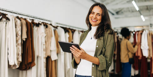 Female clothing store owner using her tablet to manage her small business with efficiency. Business woman smiling at the camera while conducting a quality check of her inventory. - JLPSF30258