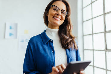 Eine kreative Geschäftsfrau arbeitet in ihrem modernen Büro und lächelt zufrieden, während sie ein Tablet benutzt, um neue und innovative Ideen zu entwickeln. Eine Frau nutzt Technologie, um ihr Unternehmen voranzubringen. - JLPSF30276