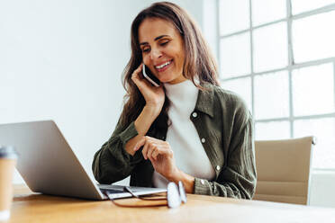 Eine Frau führt ein Telefongespräch mit ihren Geschäftskollegen in einem Büro. Eine Unternehmerin lächelt, als sie Pläne für eine Zusammenarbeit am Telefon bestätigt. - JLPSF30288