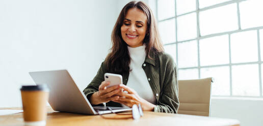 Geschäftsfrau, die lächelnd eine Textnachricht von ihren Geschäftskontakten auf ihrem Smartphone liest. Unternehmerin, die ein erfolgreiches Startup in einem modernen Büro leitet. - JLPSF30289