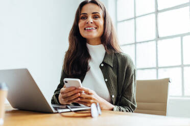 Successful startup owner smiling at the camera in her office. Happy business woman using a mobile phone as she works at her desk, using mobile apps for the day to day running of her business. - JLPSF30290