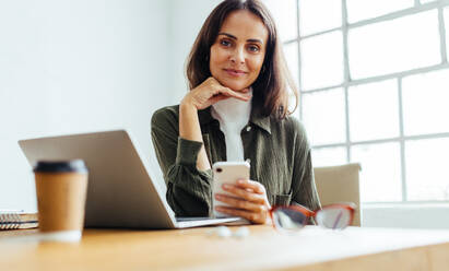 Female entrepreneur looking at the camera with confidence as she sits at her desk with a smartphone in her hand. Professional business woman using technology to run her startup. - JLPSF30292