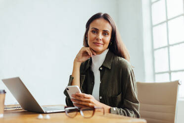 Business woman looking at the camera while sitting at a desk in her office, holding a mobile phone in her hand. Professional woman running her own startup. - JLPSF30294