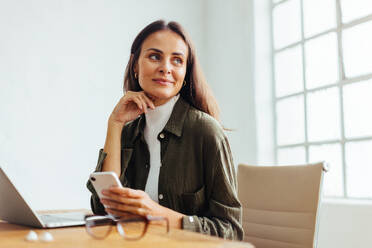 Thoughtful business woman sitting in an office with a smartphone in her hand and a laptop on her desk. Professional woman working with mobile technology in her startup. - JLPSF30295