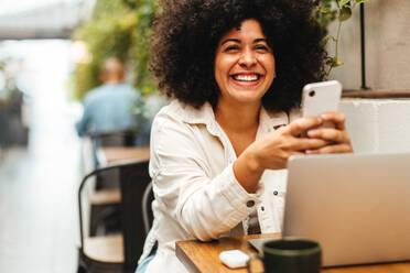 Eine afrohaarige Frau sitzt in einem Café und benutzt ihr Mobiltelefon, um online zu bleiben. Eine glückliche junge Frau genießt eine entspannende Kaffeepause mit drahtloser Technologie. - JLPSF30297