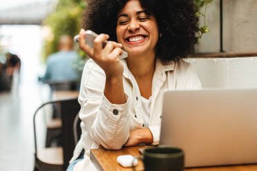 Glückliche Frau mit lockigem Afrohaar, die in einem Coworking Space arbeitet, an einem Tisch sitzt und lächelt, während sie mit ihrem Handy telefoniert. Frau, die in einem Café ihre freiberufliche Tätigkeit ausübt. - JLPSF30299