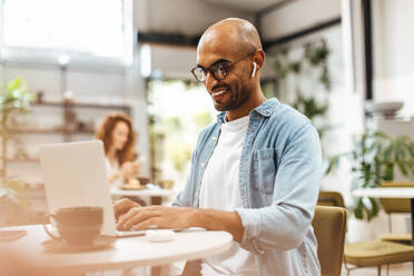 Ein Doktorand arbeitet in einem Restaurant an einem Laptop und nutzt die ruhige Atmosphäre, um sich auf sein Studium zu konzentrieren. Ein glücklicher Mann recherchiert in einem Café online für seine Diplomarbeit. - JLPSF30319