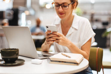 Eine Geschäftsfrau nimmt sich einen Moment Zeit, um eine Textnachricht auf ihrem Telefon zu lesen, während sie von einem Café aus arbeitet und ihre Kommunikation und Aufgaben mit Gelassenheit und Effizienz erledigt. - JLPSF30324