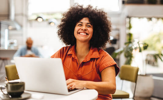 Weibliche digitale Nomadin lächelt glücklich, während sie in einem Kaffeehaus an ihrem Laptop tippt. Afrohaarige Frau in Freizeitkleidung, die in einer entspannten Umgebung online arbeitet. - JLPSF30325