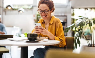 Zufriedene Studentin, die eine Kaffeepause in einem Café genießt und eine Textnachricht auf ihrem Telefon liest. Eine Studentin lächelt, während sie sich mit ihren Freunden über Online-Nachrichten austauscht. - JLPSF30326