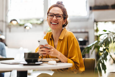 Professionelle Frau, die in die Kamera lächelt, während sie ihr Mobiltelefon in einem Restaurant benutzt, um mit Kollegen und Kunden zu kommunizieren. Glückliche junge Geschäftsfrau, die aus der Ferne arbeitet. - JLPSF30327