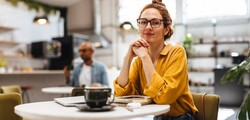 Portrait of a young female entrepreneur sitting in a cafe, looking at the camera with eyeglasses. Caucasian, ginger-haired woman doing remote work in a trendy restaurant. - JLPSF30328