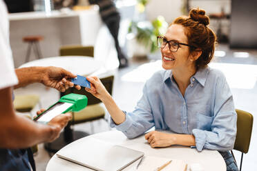 Weibliche Kundin, die mit ihrer Kreditkarte elektronisch bezahlt und die Rechnung in einem Café begleicht. Glückliche rothaarige Frau, die von einem Kellner in einem Restaurant bargeldlos bedient wird. - JLPSF30334