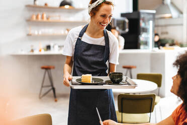 Professionelle und einladende Barista, die einer treuen Kundin Kaffee und Kuchen auf einem Tablett serviert und dabei lächelt, während sie das Essen ausliefert. Café-Kellnerin, die einer Kundin einen hochwertigen Service bietet. - JLPSF30342