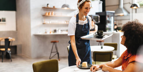 Eine Restaurantkellnerin lächelt und grüßt ihre Kundin freundlich, während sie ihr Essen auf einem Tablett bringt. Eine Barista serviert einer Frau eine Tasse Kaffee und ein Stück Kuchen in einem Café. - JLPSF30343