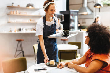 Eine Kellnerin in einem Restaurant begrüßt ihre Kunden mit einem Lächeln und bringt das Essen auf einem Tablett. Eine freundliche Barista serviert einem Kunden eine Tasse Kaffee und ein Stück Kuchen. - JLPSF30344