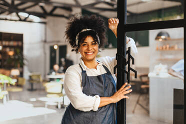 Cafe owner stands inside her small business, wearing an apron and beaming with a smile. Happy young entrepreneur ready to serve her customers with delicious drinks and a warm dining experience. - JLPSF30349