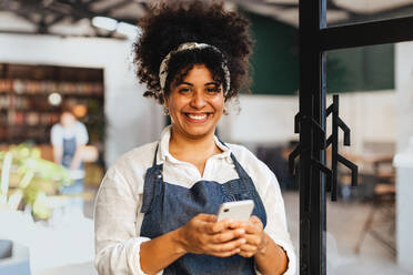 Eine glückliche junge Frau und Restaurantbesitzerin hält ihr Telefon in der Hand und nutzt gekonnt die mobile Technologie, um ihr kleines Unternehmen mit Leichtigkeit zu führen. Eine Unternehmerin, die im Gastgewerbe erfolgreich ist. - JLPSF30350
