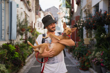 Positiver männlicher Besitzer in Freizeitkleidung und Panamahut, der auf einer schmalen Straße zwischen kleinen, mit Blumen geschmückten Wohnhäusern steht und einen liebenswerten Basenji-Hund im Arm hält - ADSF43947