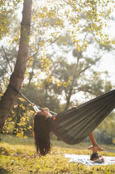 Side view of unrecognizable female with long brown hair chilling in hammock on sunny day in forest of Asturias - ADSF43950