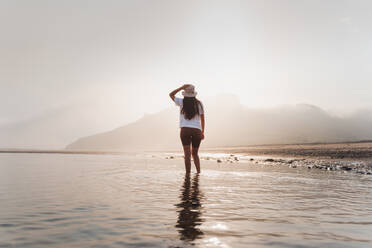 Rückenansicht einer nicht erkennbaren Frau in Sommerkleidung und Panamahut, die an einem sonnigen Tag in Asturien im Meerwasser nahe der Küste gegen einen Berg läuft - ADSF43953