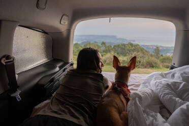 Rückenansicht einer nicht erkennbaren weiblichen Reisenden, die auf einer bequemen Matratze neben einem gehorsamen Basenji-Hund liegt und die malerische Landschaft Asturiens bewundert - ADSF43954